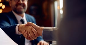 A business owner and a fiduciary wealth advisor shaking hands across a desk after an M&A consultation.